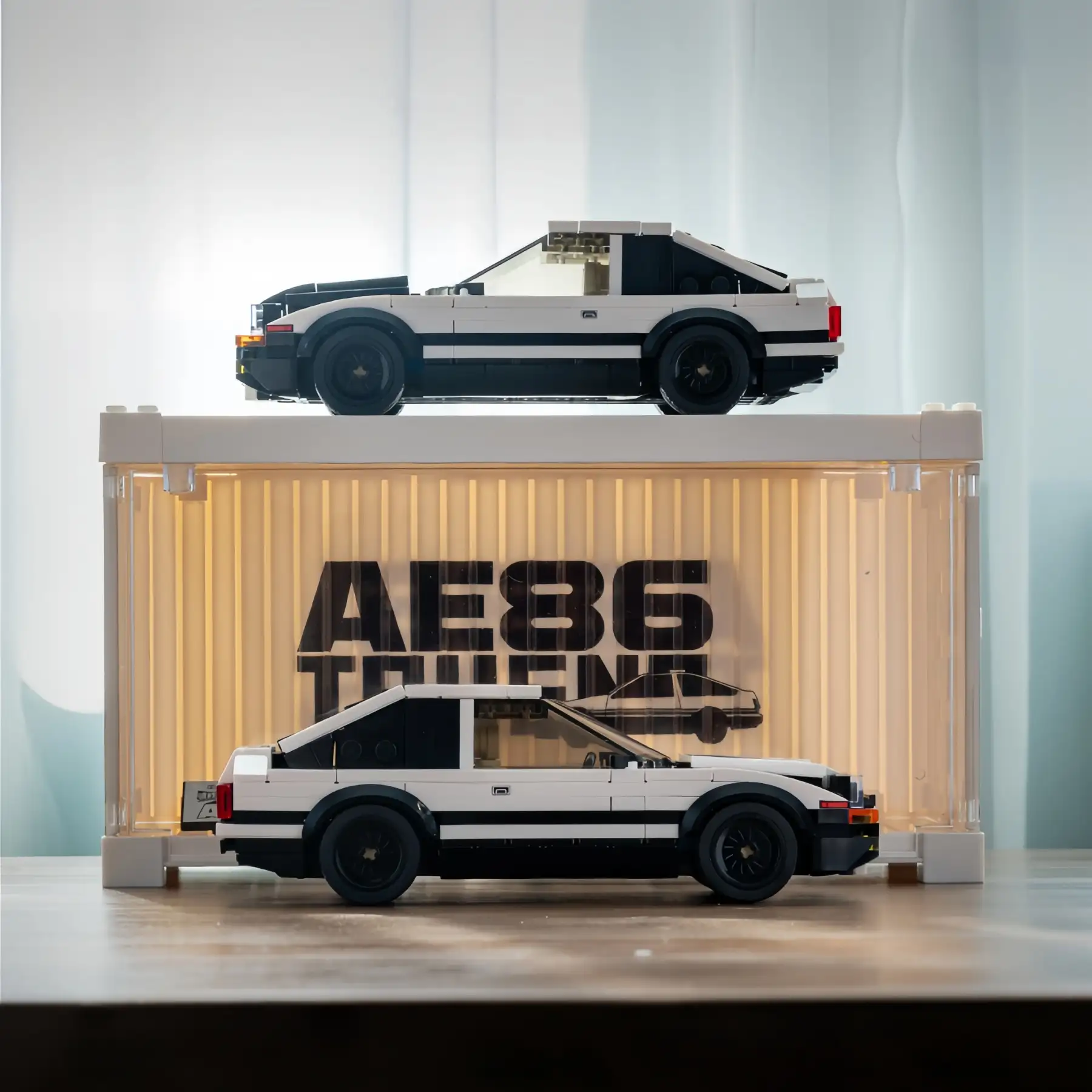 Two Toyota AE86 Trueno brick model cars displayed with an illuminated white shipping container display box on a table.