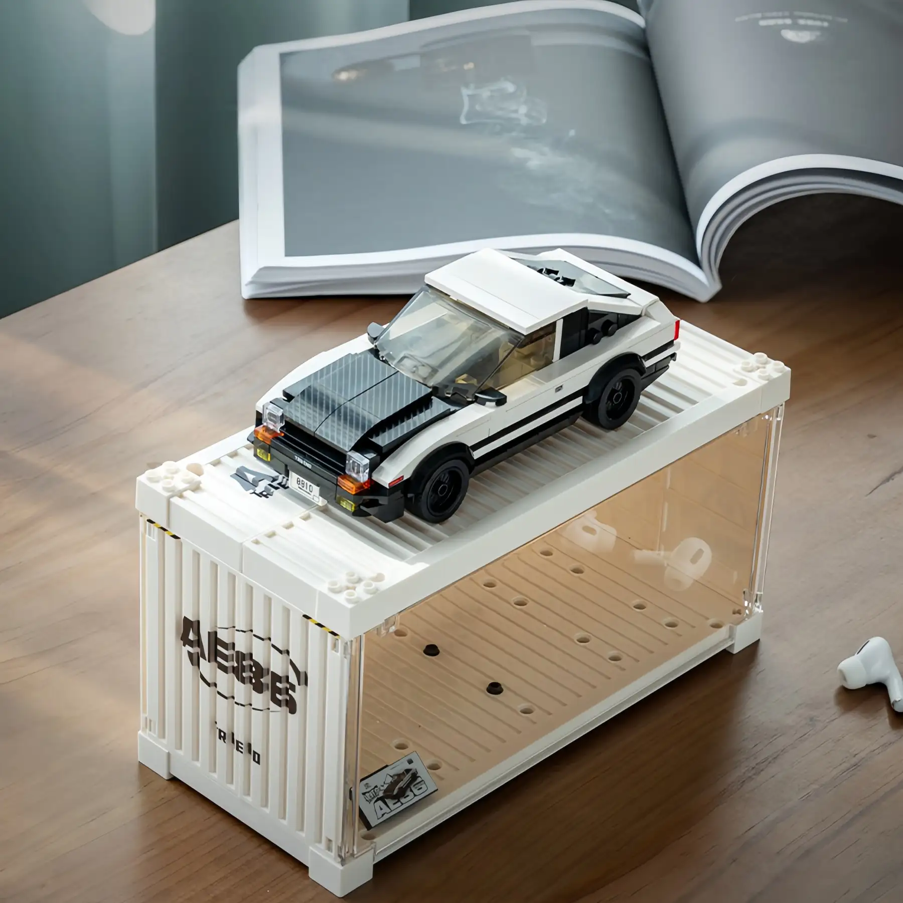 Toyota AE86 brick car parked on top of its white shipping container display stand on a wooden table.